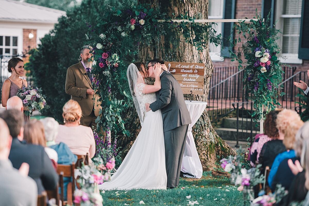 Bride wearing a long white dress and veil kisses the groom in a gray suite at Rippavilla Mansion. Their lush arbor of greenery, pink, white and purple flowers.