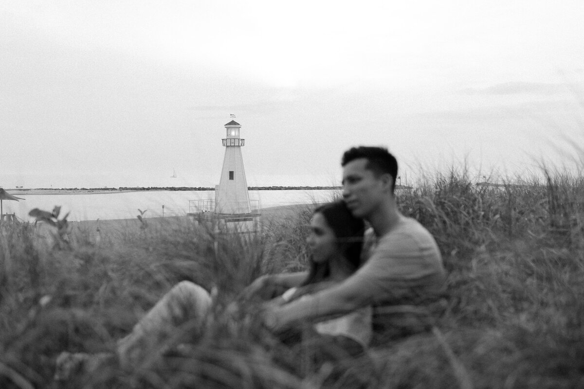 Woman sits leaning back on man in long grass at the beach in front of a lighthouse