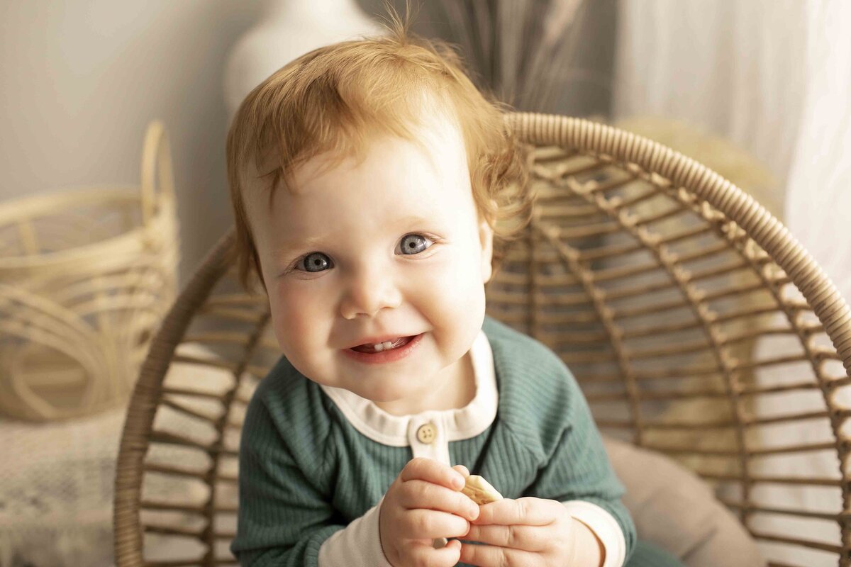 One-year-old birthday boy smiling and sitting comfortably in a wicker chair, surrounded by soft studio lighting