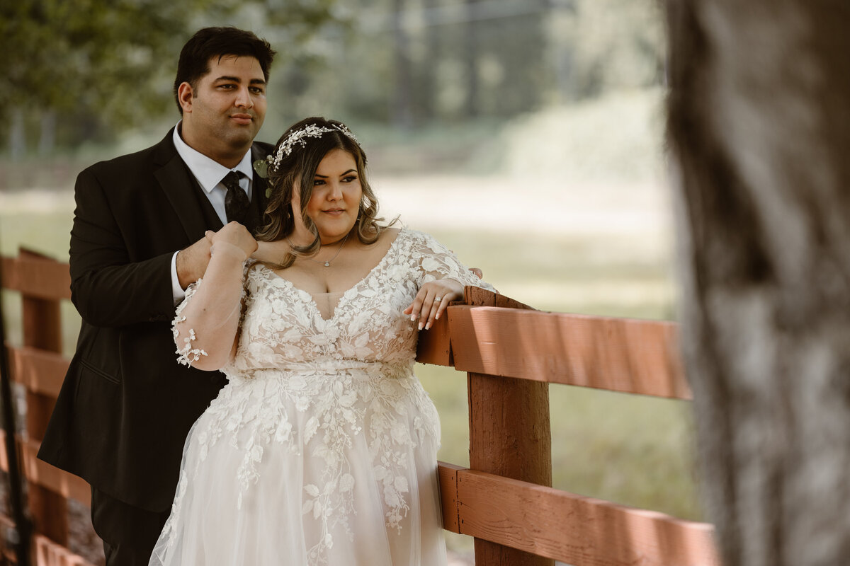 Bride and groom stand closely together against a wooden ranch fence, bathed in soft summer light, with the bride in a lace gown and the groom in a black suit.