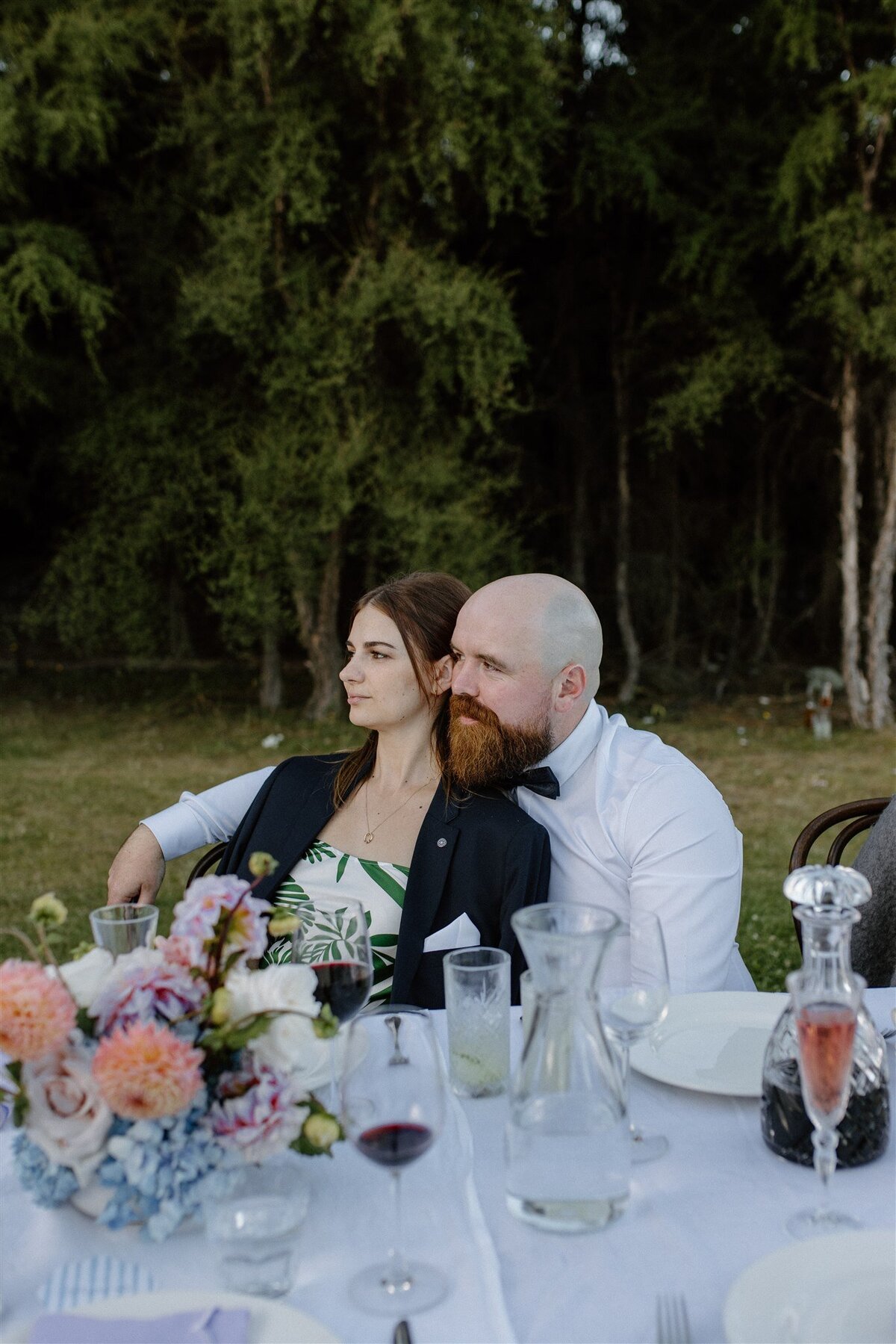 A couple cuddled together at an alfresco wedding reception table, surrounded by florals, glassware, and soft evening light with a forest backdrop.