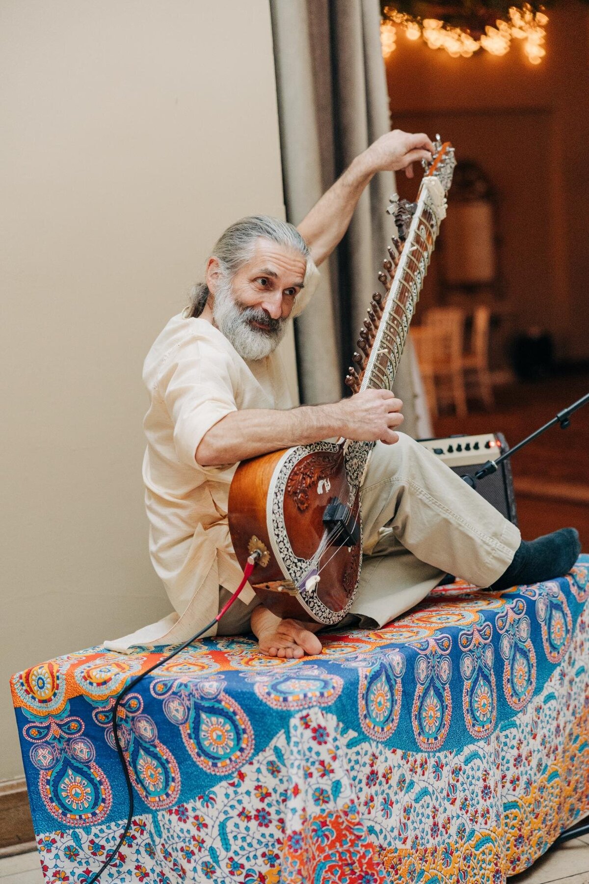 An elderly man with a long beard tuning a sitar while sitting on a colorful patterned cloth-draped platform.