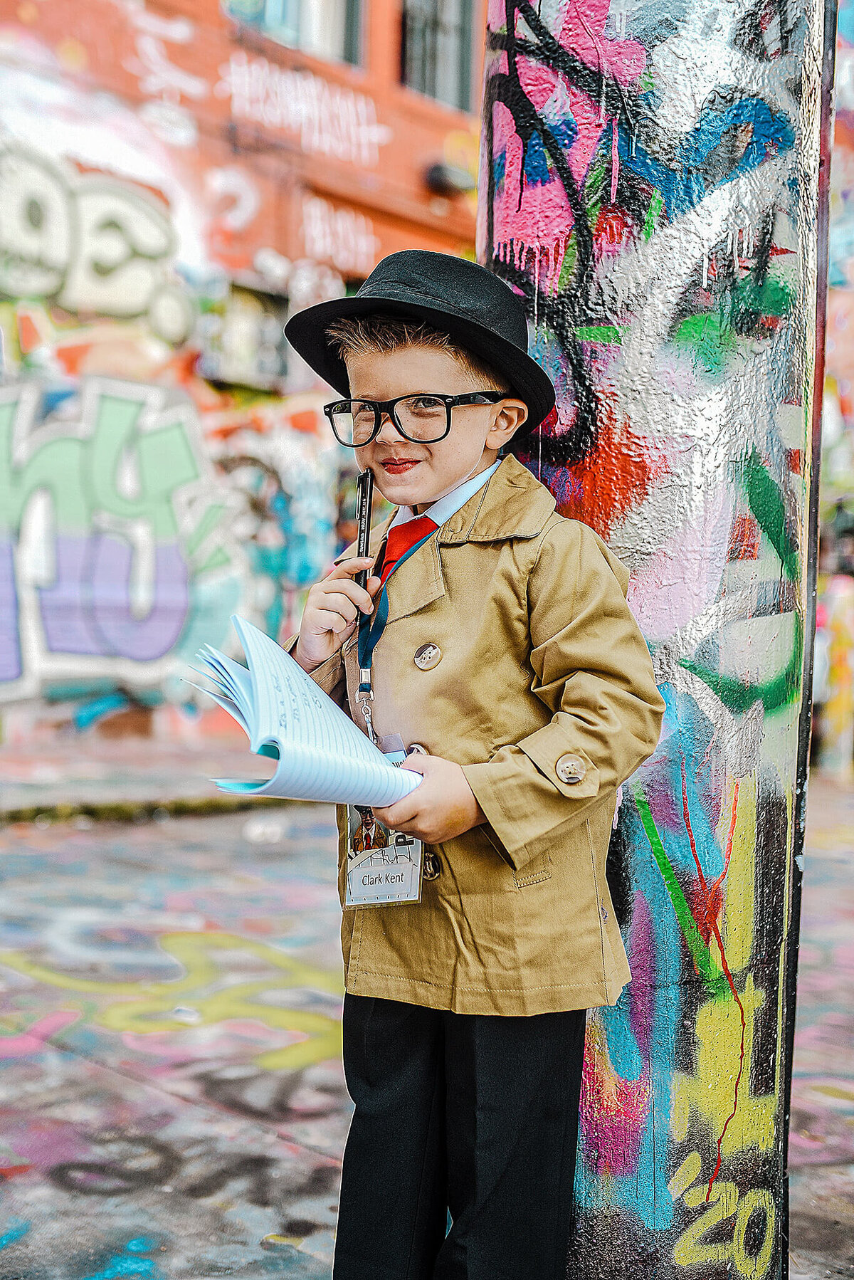 Little boy with black fedora hat holding a pen in a trench coat at graffiti alley in Baltimore Maryland near MICA