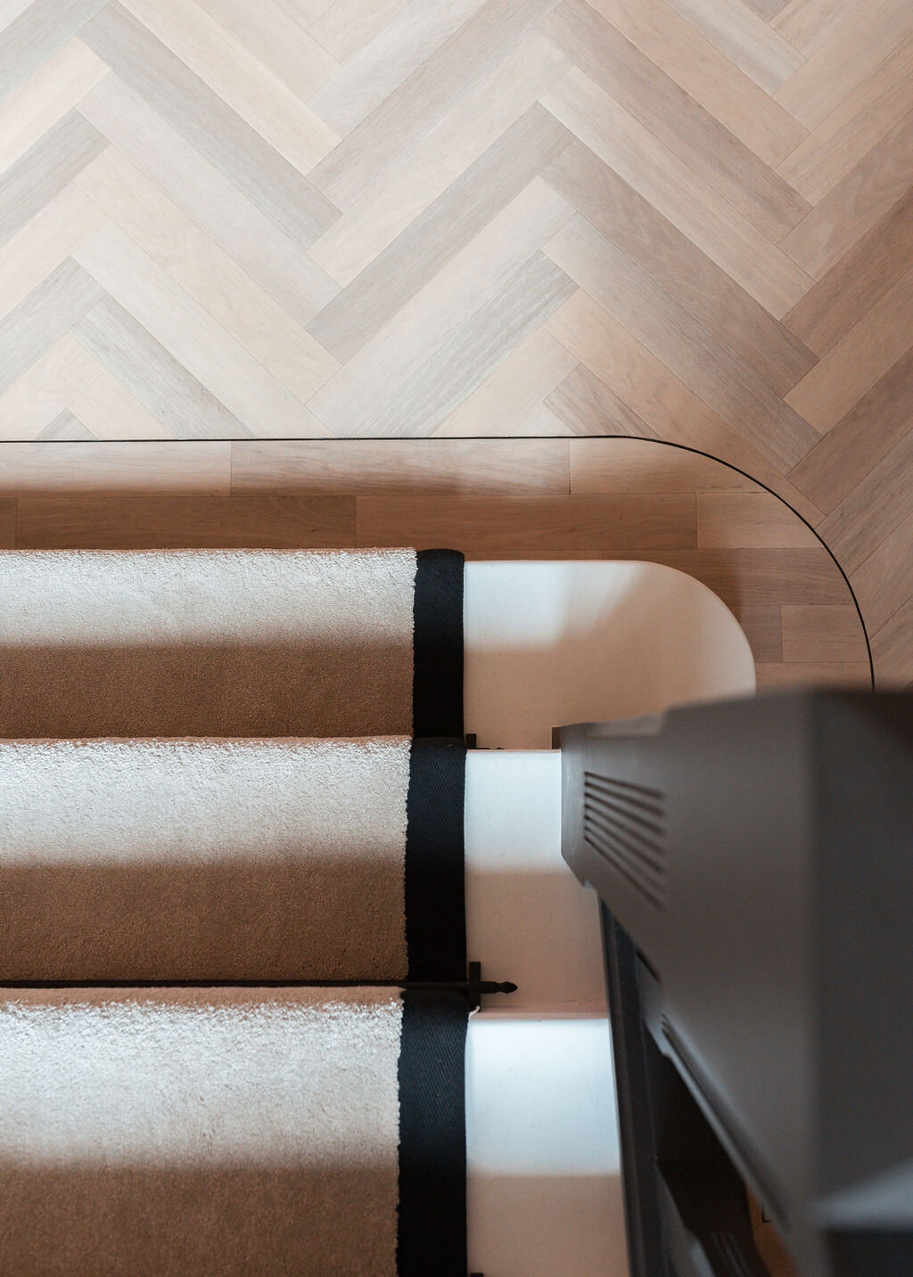 Aerial view of a white staircase featuring a beige runner bordered by black trim. Surrounding the staircase is light wood flooring laid in a herringbone pattern. The top of a black banister is partially visible in the lower right corner.