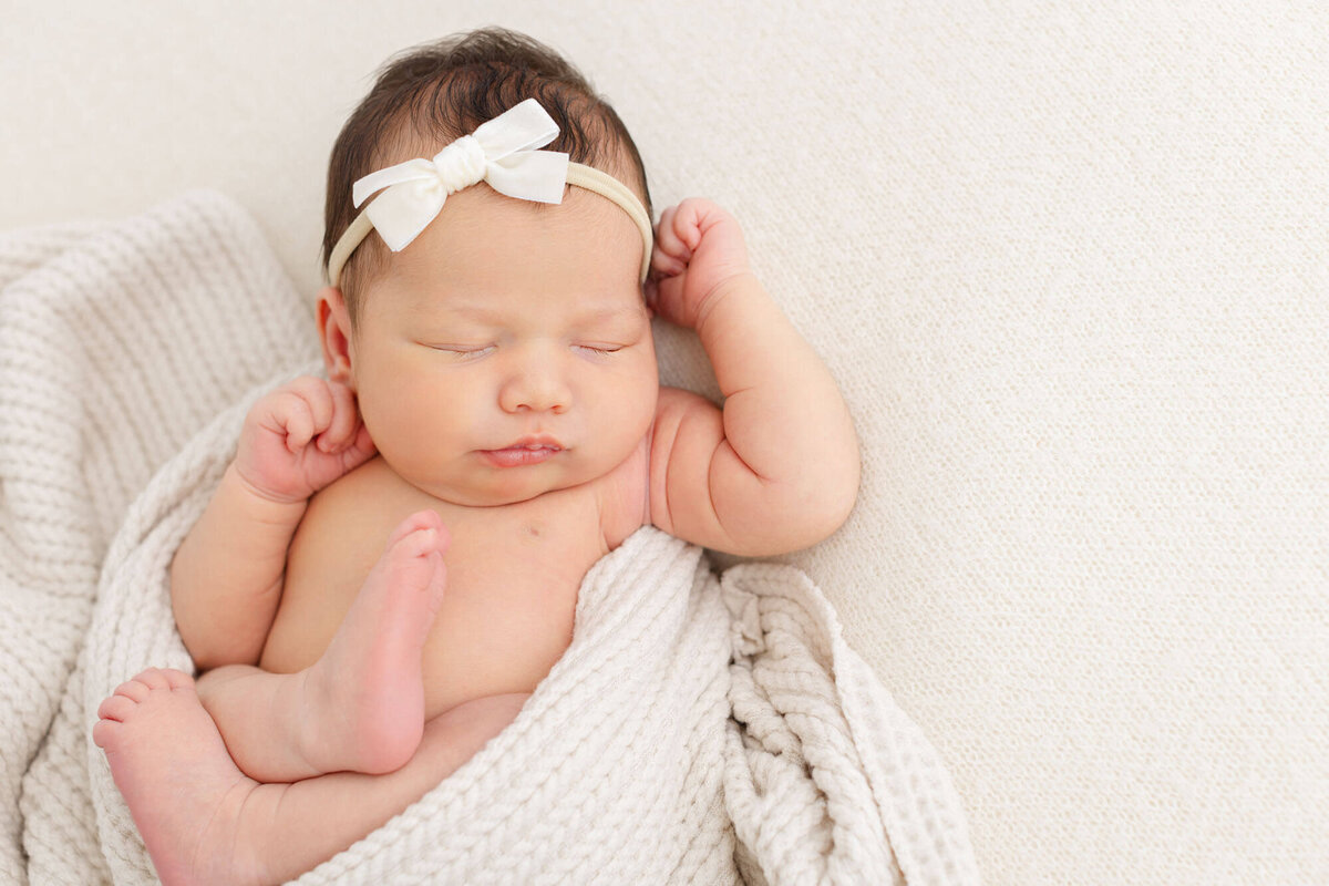 baby wrapped in a textured beige blanket with feet and arms out and a white bow on her head. Newborn baby is sleeping sweetly at her Portland newborn photoshoot.