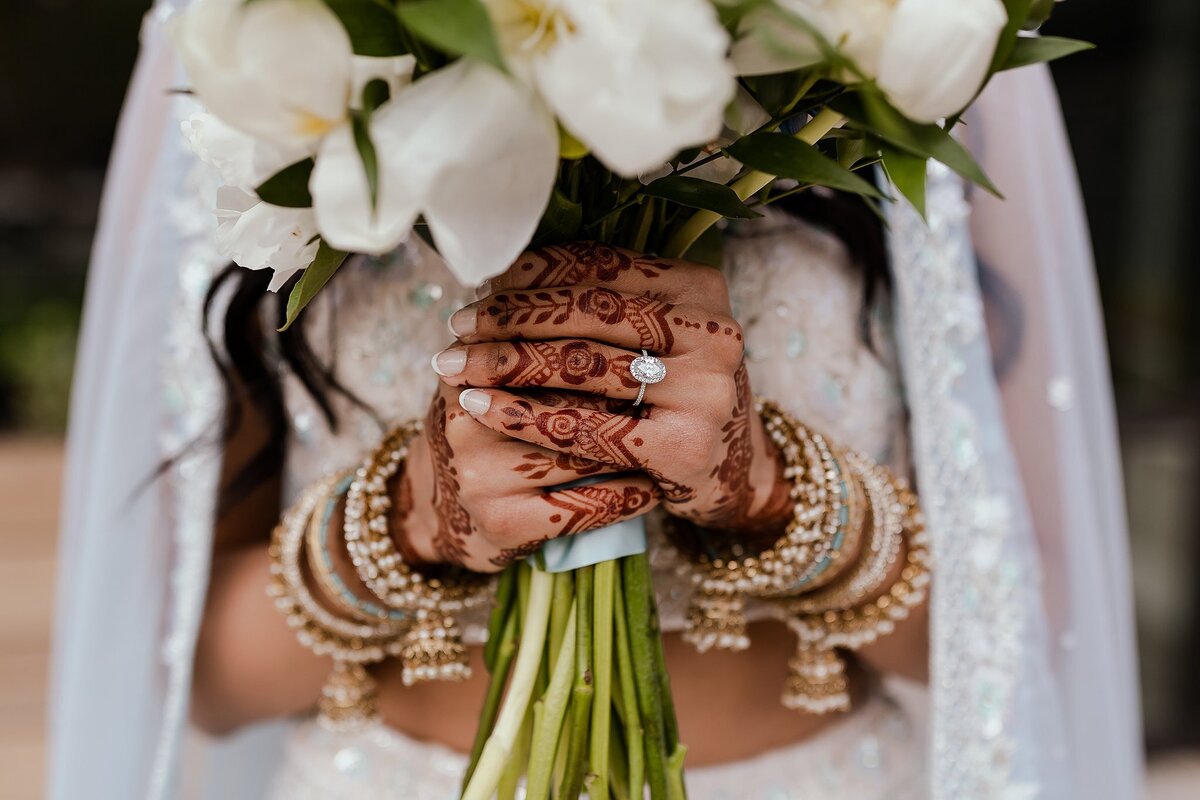 Indian bride holds a large white and green bridal bouquet displaying her Indian wedding jewelry and her mendhi. The Hindu bride is wearing a light blue dupatta and an oval cut diamond engagement ring.