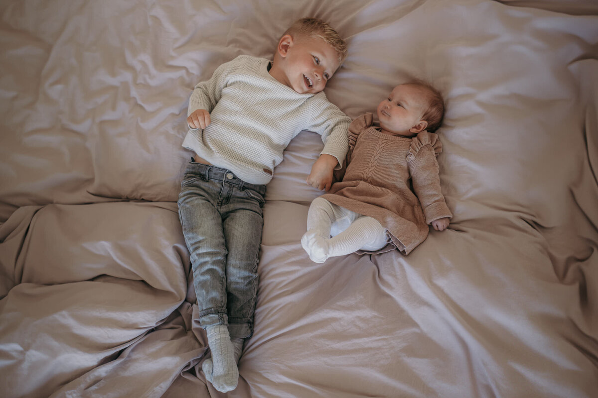Siblings laying on a bed, holding hands.