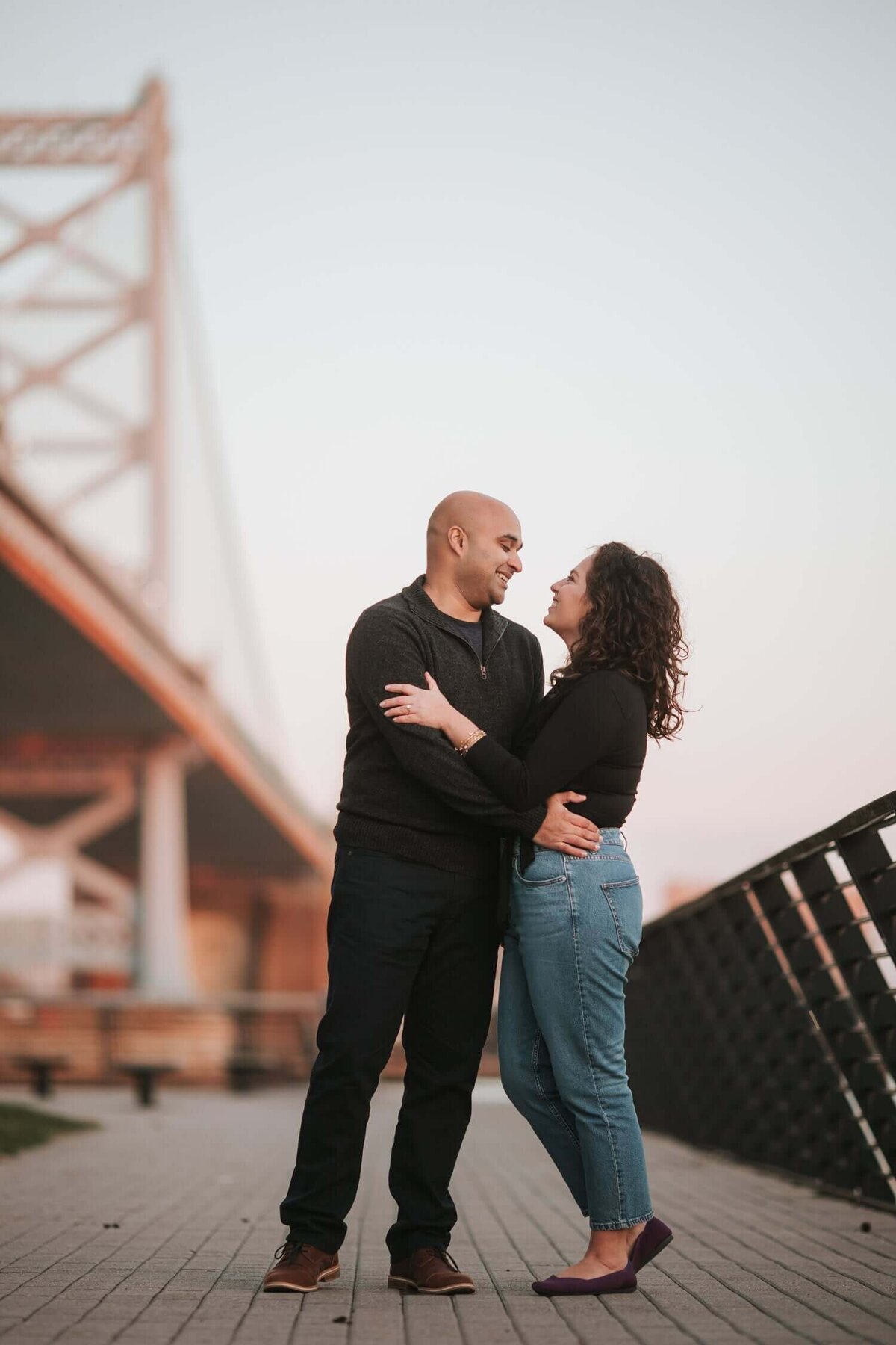 A soon-to-be wedded couple standing next to a bridge embracing each other in Philadelphia.