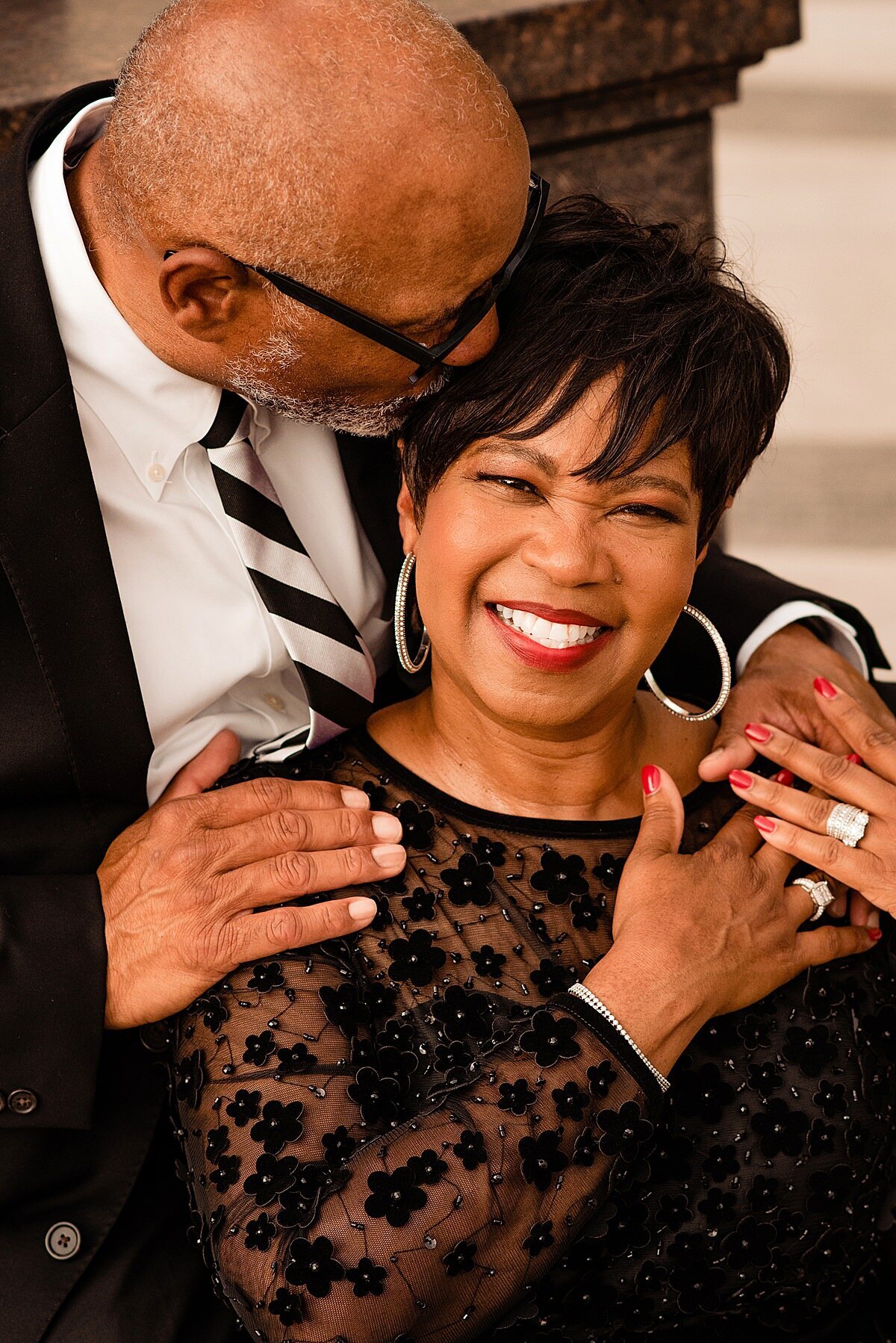 The groom embraces the bride from behind as he kisses her temple. The groom is wearing a black suit with a white shirt and a black and white striped tie. The bride is smiling up at the camera.  She is wearing large silver hoop earrings and a long sleeved sheer black dress with small black flowers all over it accented with tiny rhinestones.