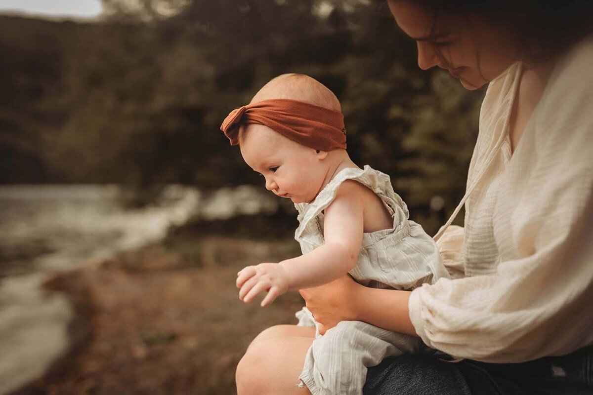 A mother holding her curious baby on her lap while they both look down at the water on the lake