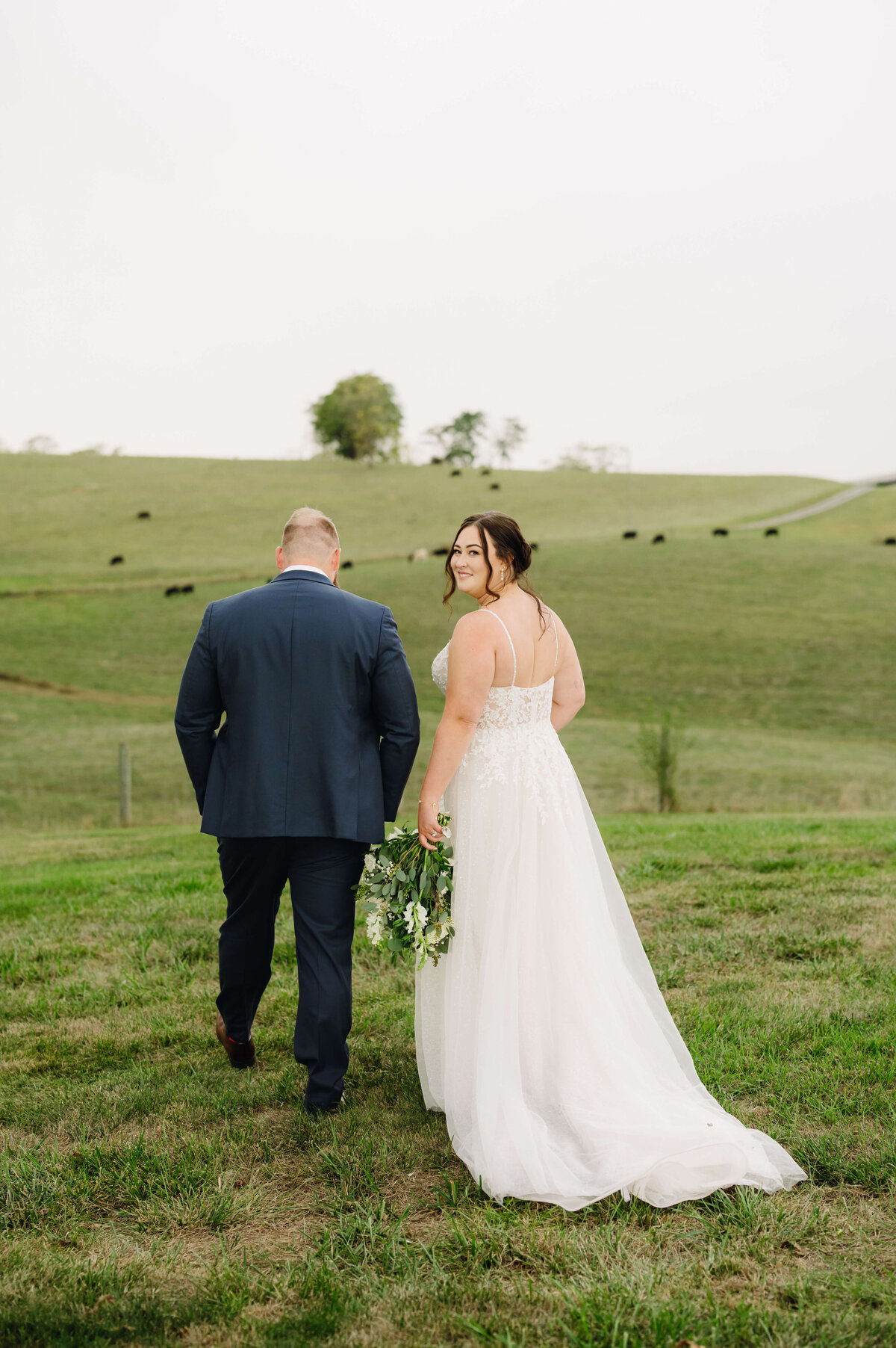 Charlottesville wedding photographer captures candid picture of bride and groom walking towards a field of cattle at Sunny Slope farm with the bride smiling over her shoulder