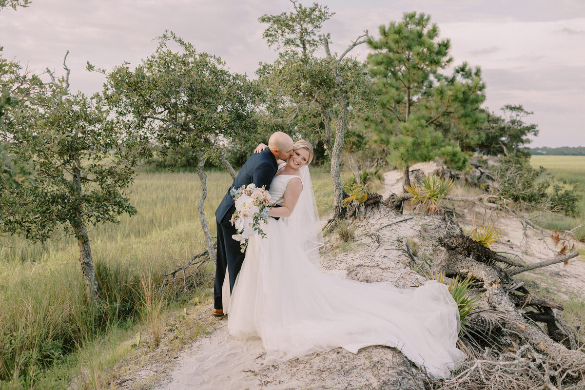 Image by Georgia wedding photography image of groom in navy tuxedo dipping bride holding a cascading bouquet of pastel florals while kissing her cheek. Her long train hangs over the side of a peak overlooking the summertime marsh with lush greens overcast sky.