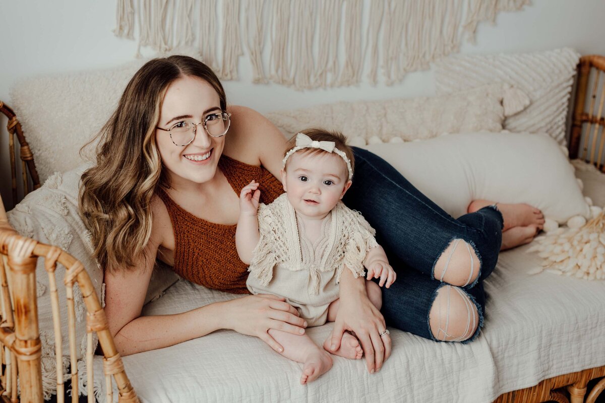 Studio lifestyle baby session - Mom laying on her side smiling on a couch and baby sitting in front of her. Mom and baby smiling at the camera. Boho vibes.