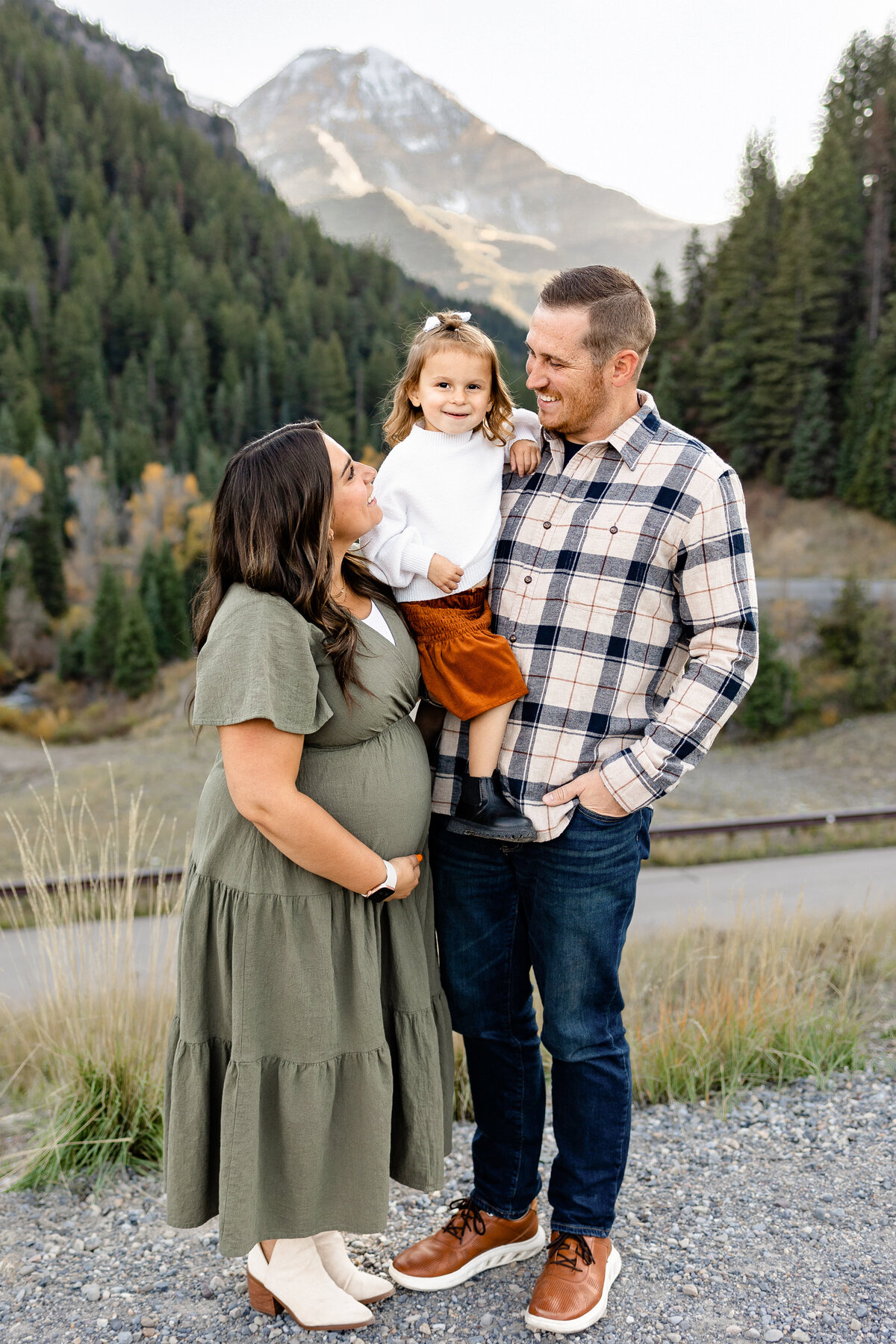 A pregnant mother in an olive-green dress, a father in a plaid shirt, and their little daughter in a white sweater with a rust-colored skirt stand together, gazing at each other with love in front of towering mountains.