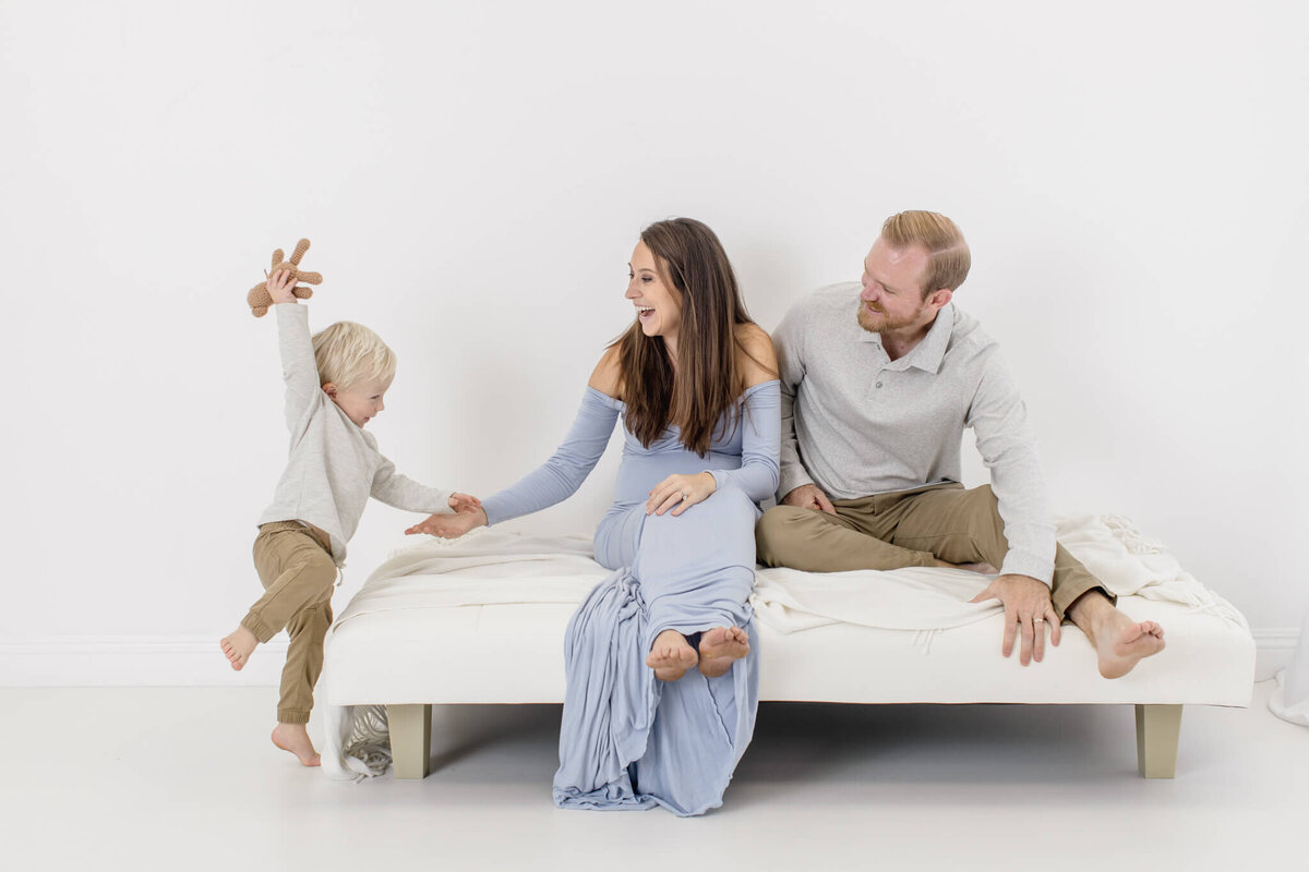 A family sitting on a bed in a bright room. The mother in a light blue dress is smiling at a young child jumping with joy. The father, wearing a light shirt and khaki pants, watches with a smile. The atmosphere is happy and playful.