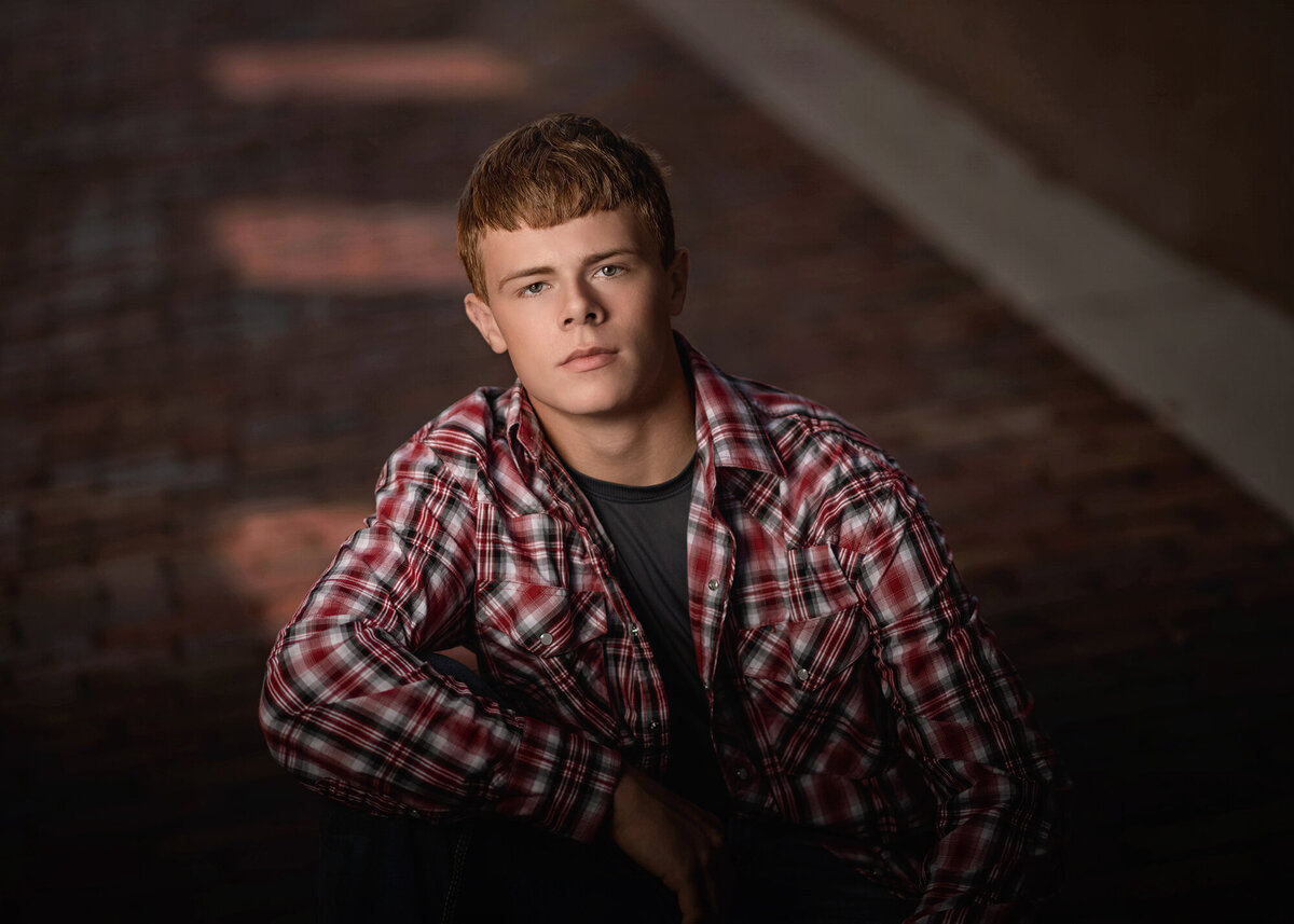 A grad leans on his knee in a brick street in a red plaid button down