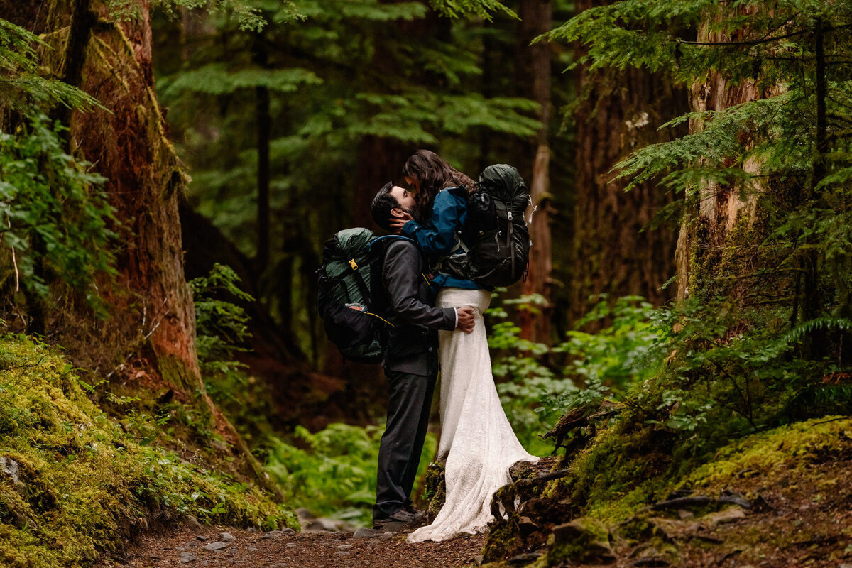 a couple in their wedding attire and hiking gear kiss on the middle of a forested path