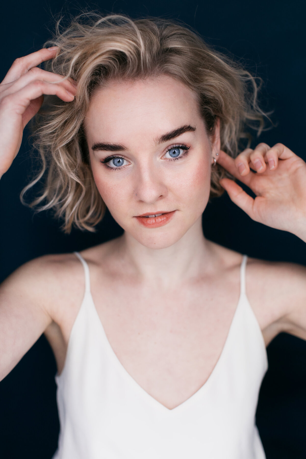 Castingfoto van Marieke Goemans. Vrouw met korte blonde krullen in een witte top voor een donkerblauwe backdrop.