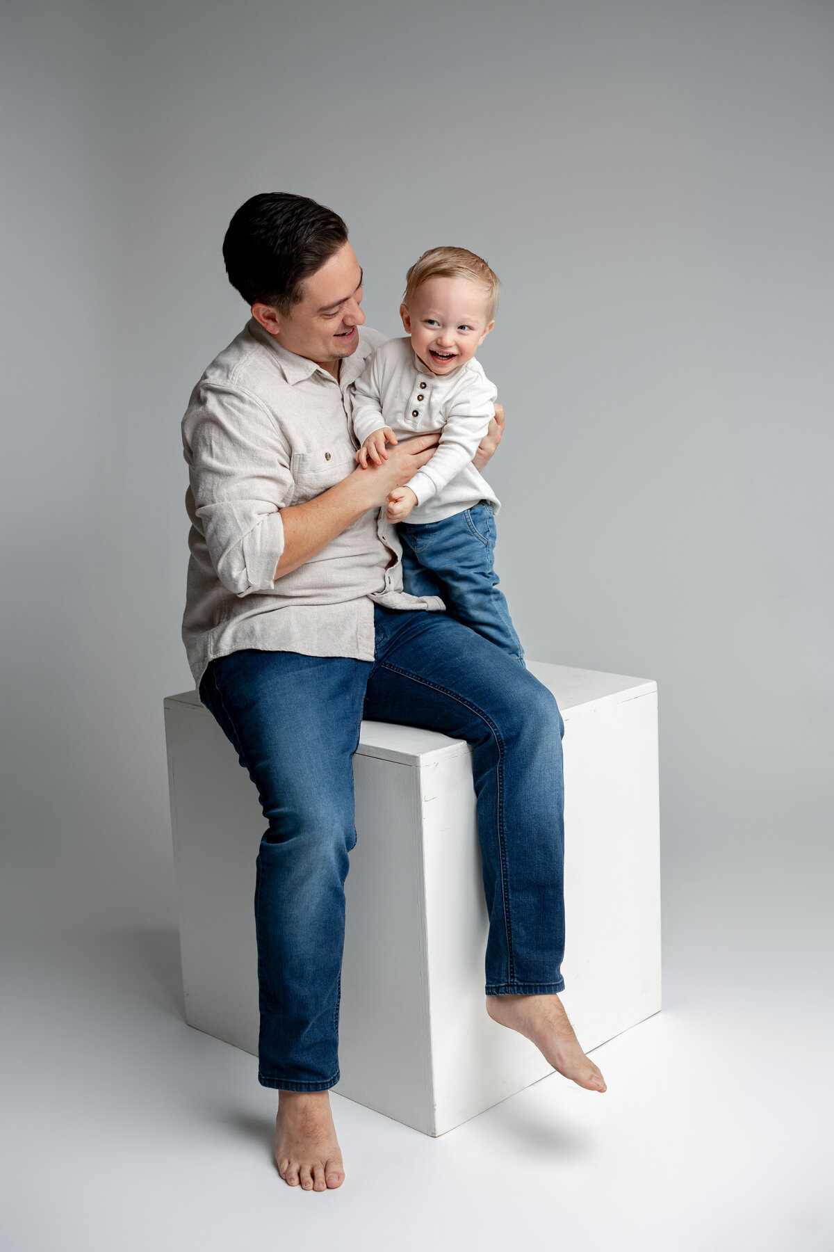 Father sitting on cube with toddler son in jeans and white shirt, smiling together. Photographed by Yucaipa family photographer Kaitlyn Dawn Photography.