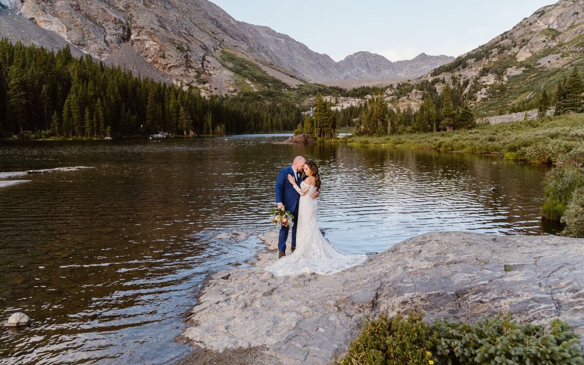 Bride and groom standing on a rocky shoreline during their Blue Lakes elopement in Breckenridge, Colorado, surrounded by alpine water, pine trees, and mountain peaks.