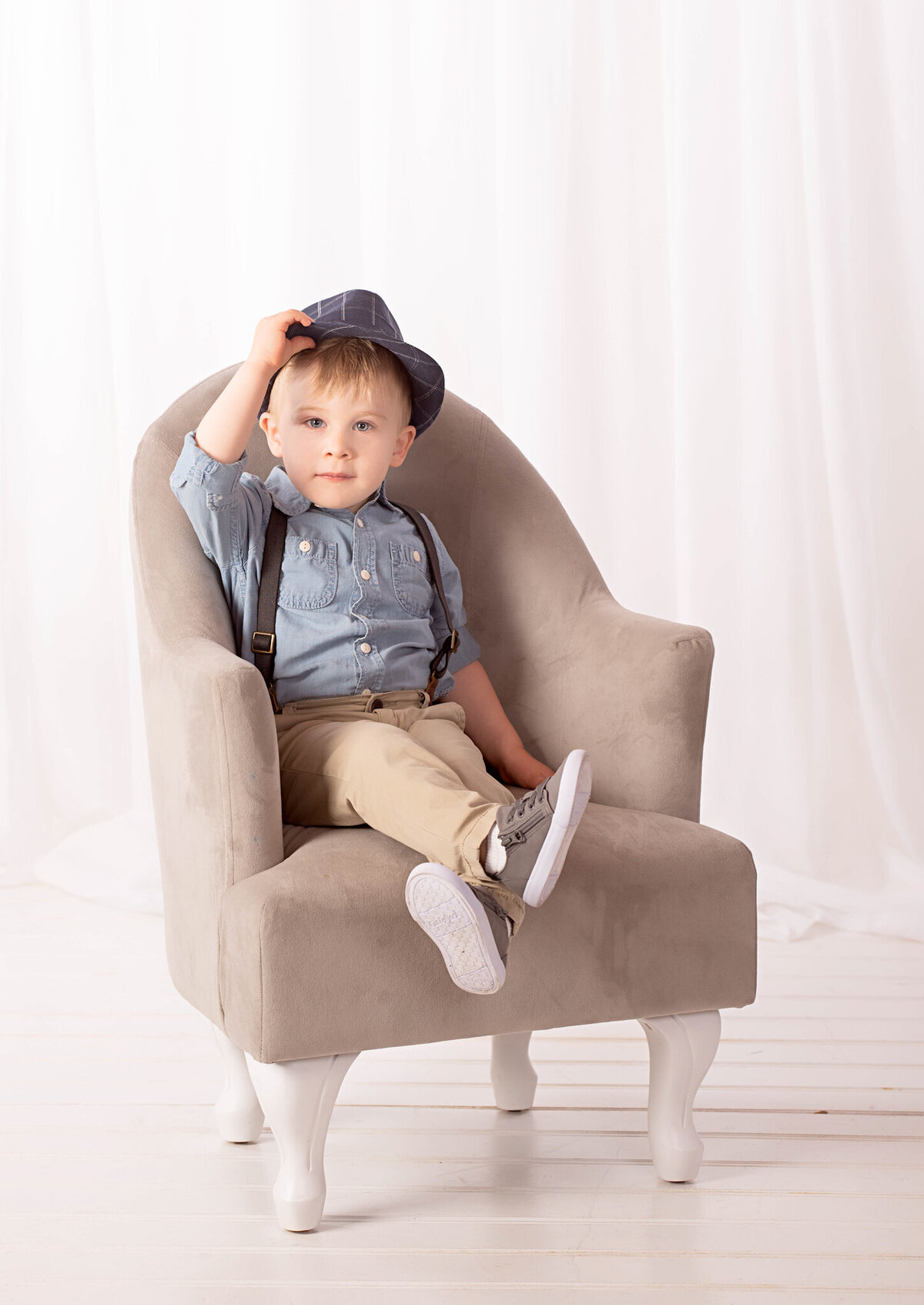 Adorable studio portrait of a toddler sitting in a modern armchair, dressed in denim and suspenders. Captured in Overland Park, Kansas, this clean and minimalist setup is perfect for milestone photography.
