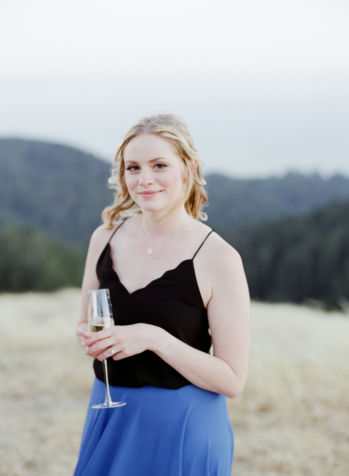 Pretty blond lady holds a glass of bubble champagne and looks compassionately at the camera. Green lush hills in the background.