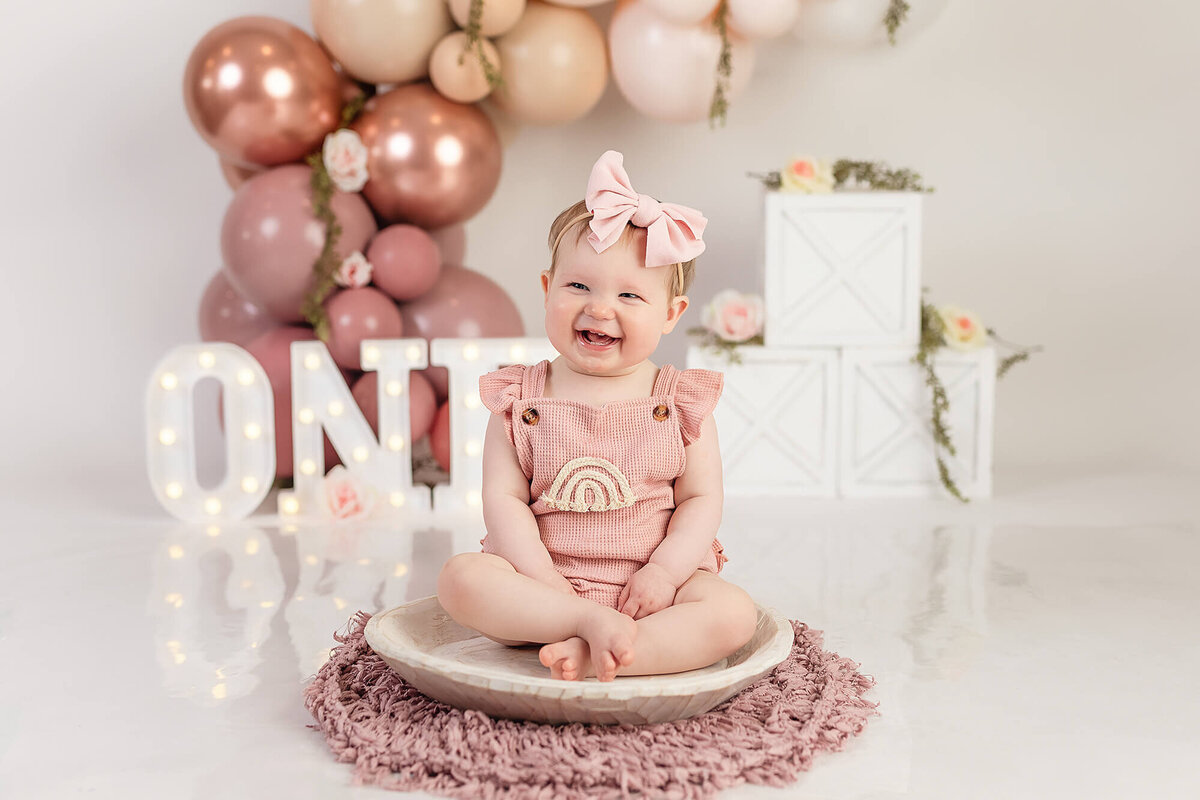 One year old girl wearing a pink rainbow romper sits in front of her balloon arch backdrop.