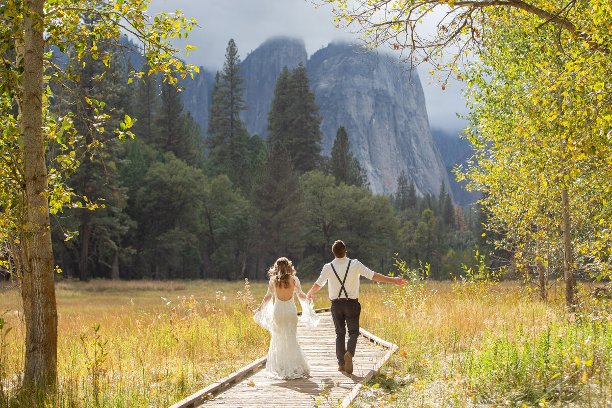 A bride and groom walk hand in hand down a wooden boardwalk in Yosemite.