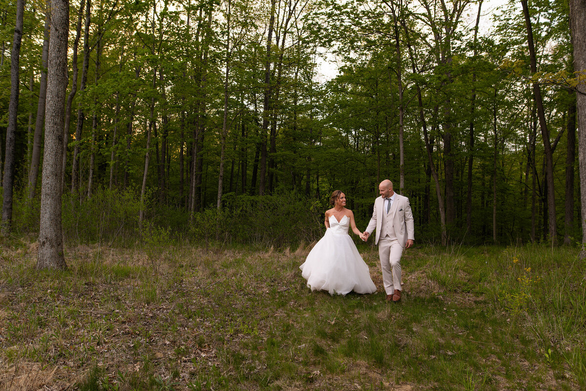 Bride and groom hold hands while walking next to a forest at sunset.