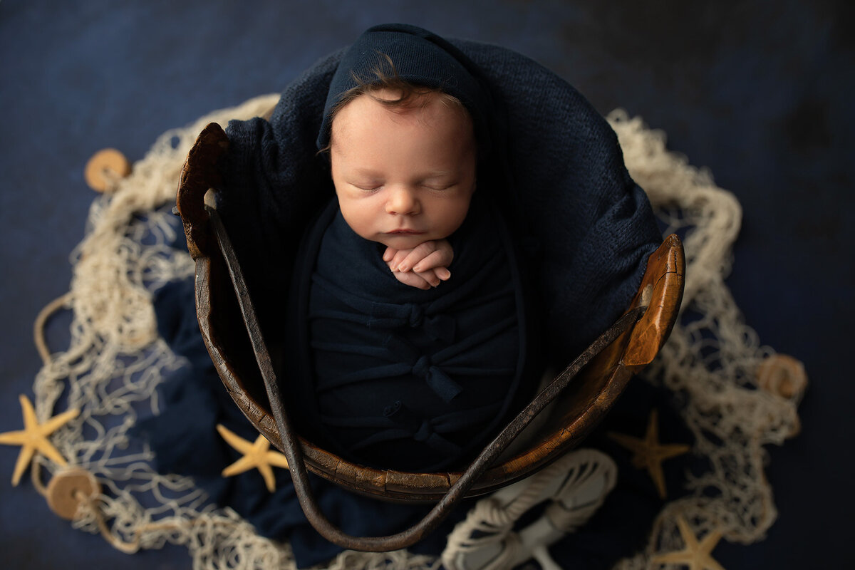 Newborn baby boy swaddled in deep navy wrap, posed in a rustic bucket with nautical props and starfish, captured by DFW newborn photographer.