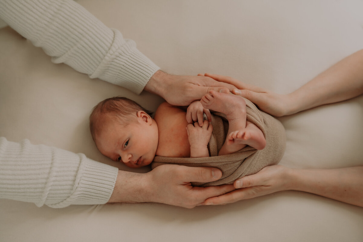 Detail foto met de handen van de ouders rond de newborn die gewikkeld is in een wrap voor zijn newbornshoot in de daglicht studio te Antwerpen.