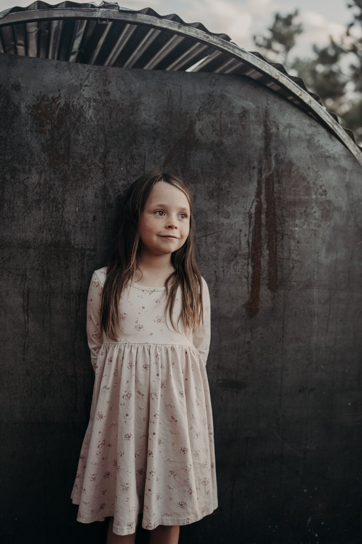 A beautiful colored portrait with a young girl, wearing a long sleeve dress instead of looking at the camera she is smiling off into the distance, with her hands, naturally held behind her.