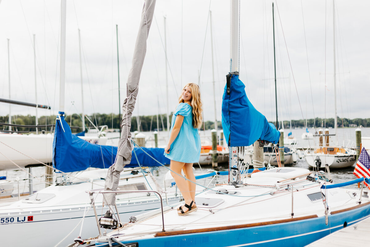 Senior girl in a blue dress standing on a sailboat with other boats in the background