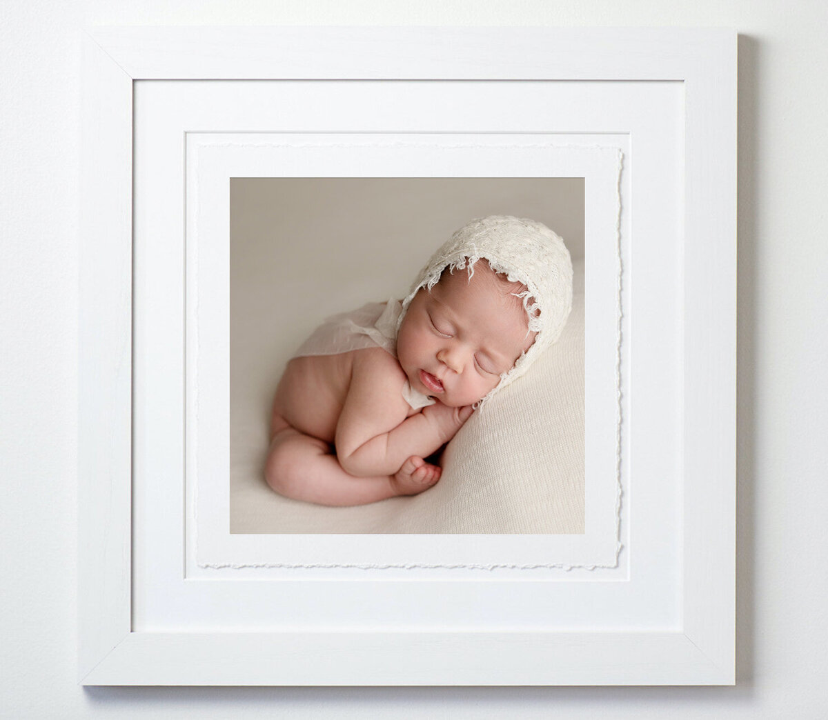 Framed deckled edge art print of a newborn baby photographed in a white bonnet on a neutral backdrop at her newborn session