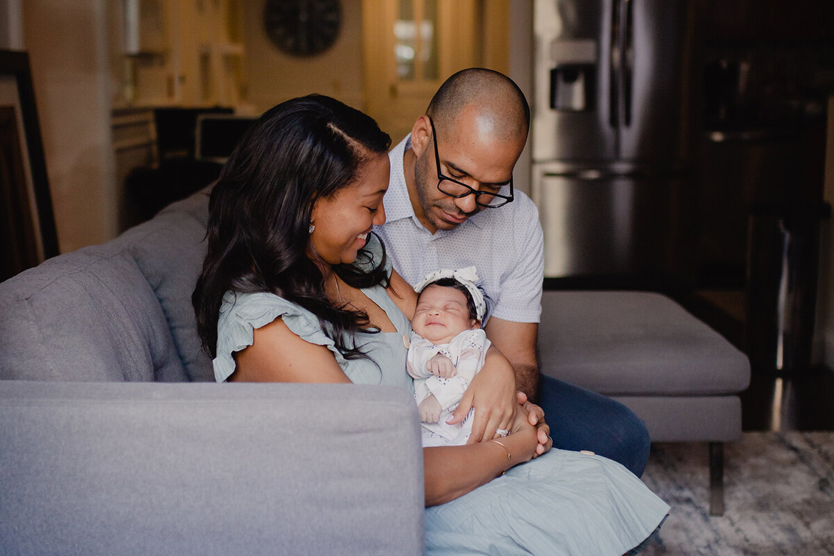 A young black family brings home their newborn baby girl during the pandemic. This photo is shot through their window into their living room. They sit on the couch and look down at their new daughter.