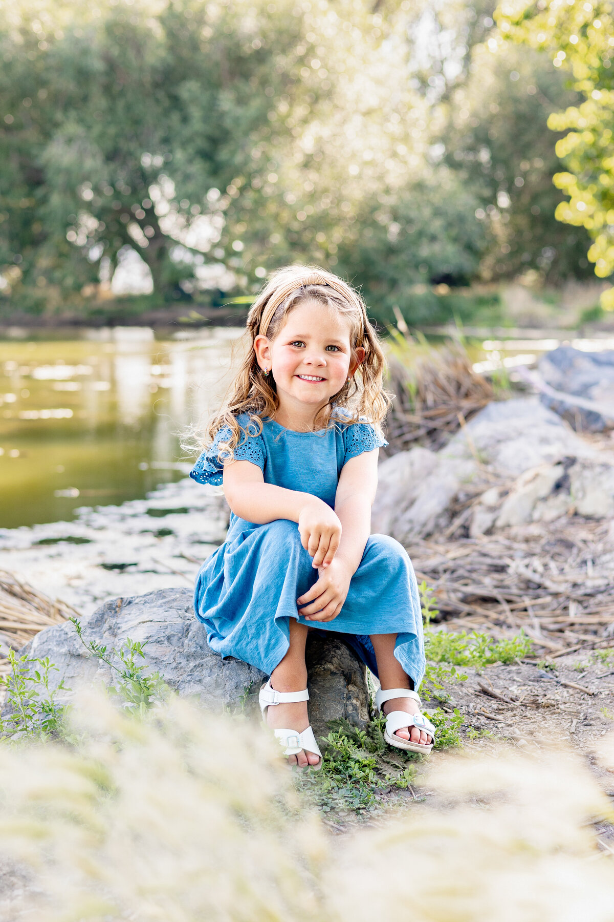 A young girl with curly blonde hair and a blue dress sits on a rock near a calm pond, smiling brightly as the sunlight filters through the trees.