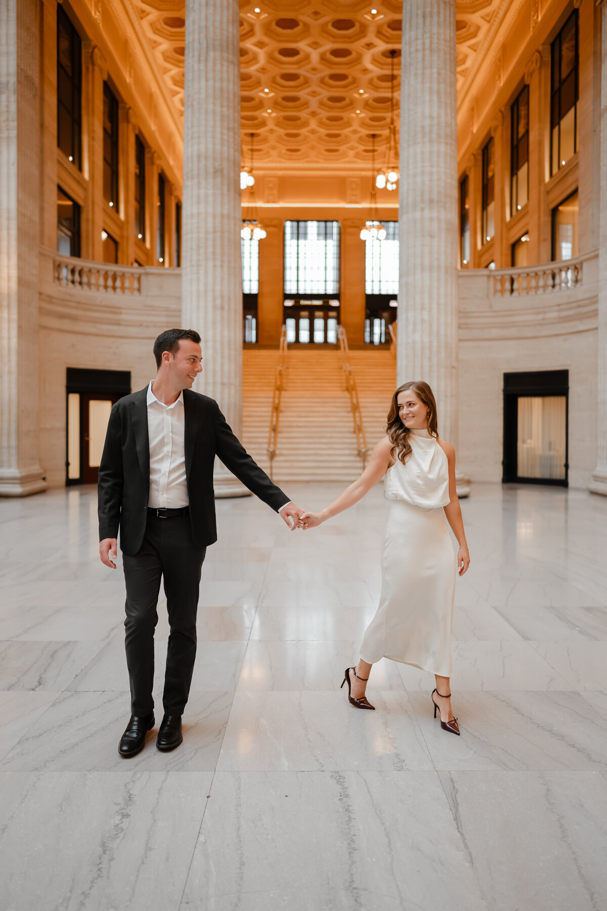 A couple hold hands while walking in front of the grand staircase at Chicago Union Station