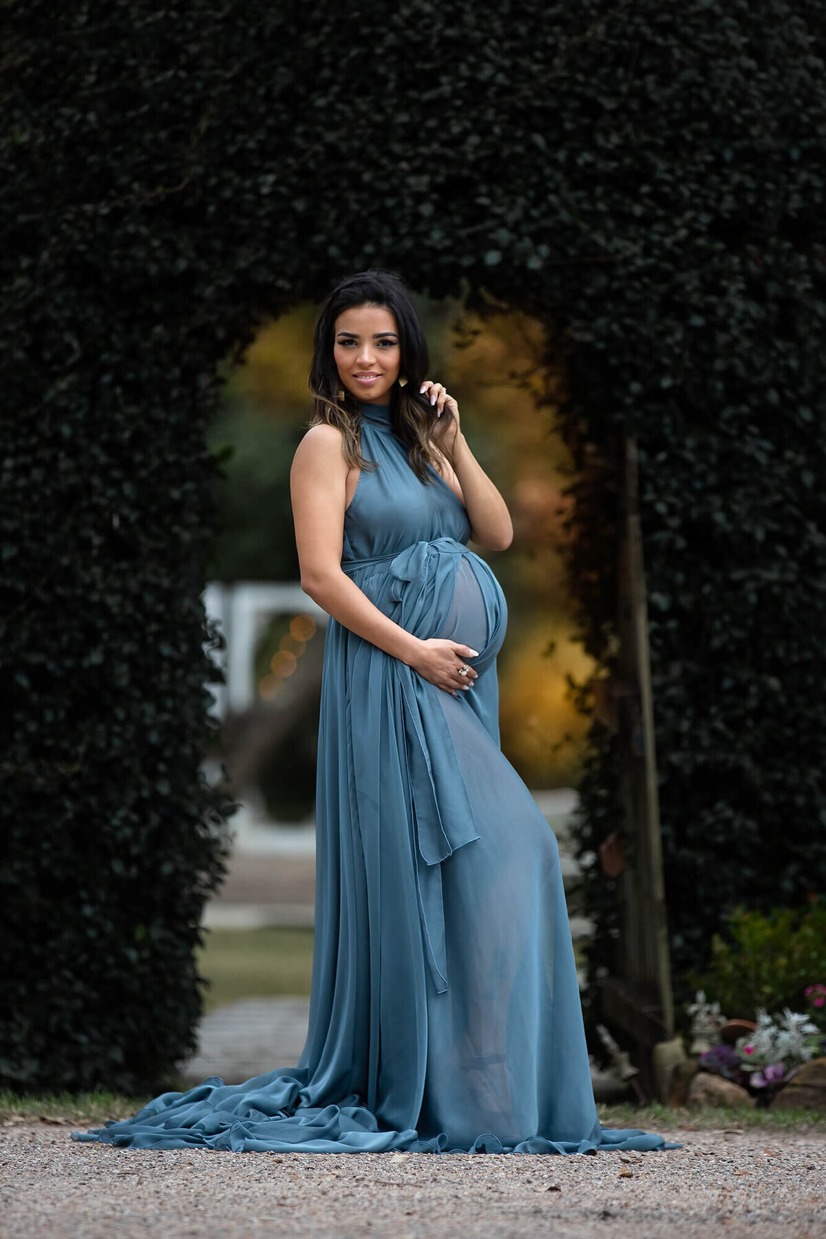 Maternity model wearing a beautiful blue dress posing under a green arch