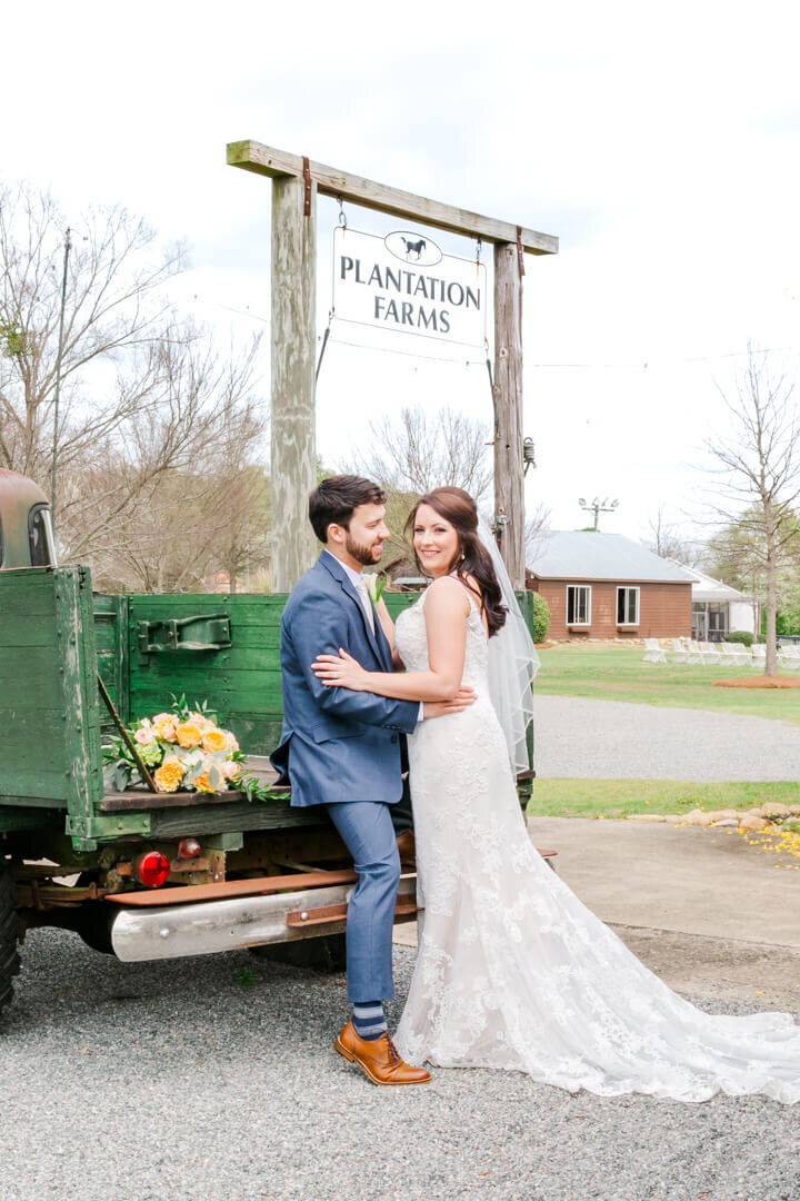 Groom sits on the back of a green truck bed while the bride stands next to him in is arms while she smiles at the camera.