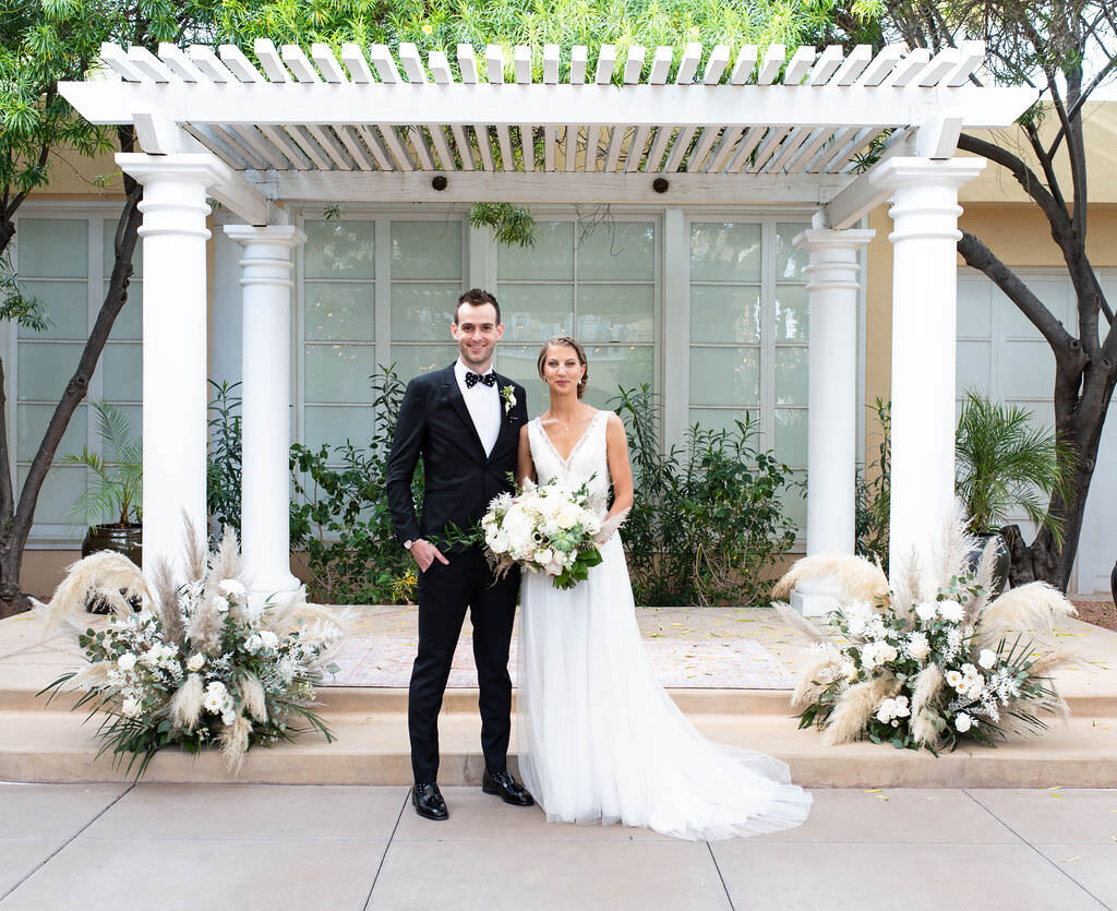 A bride and groom standing under a small awning with florals next to them.