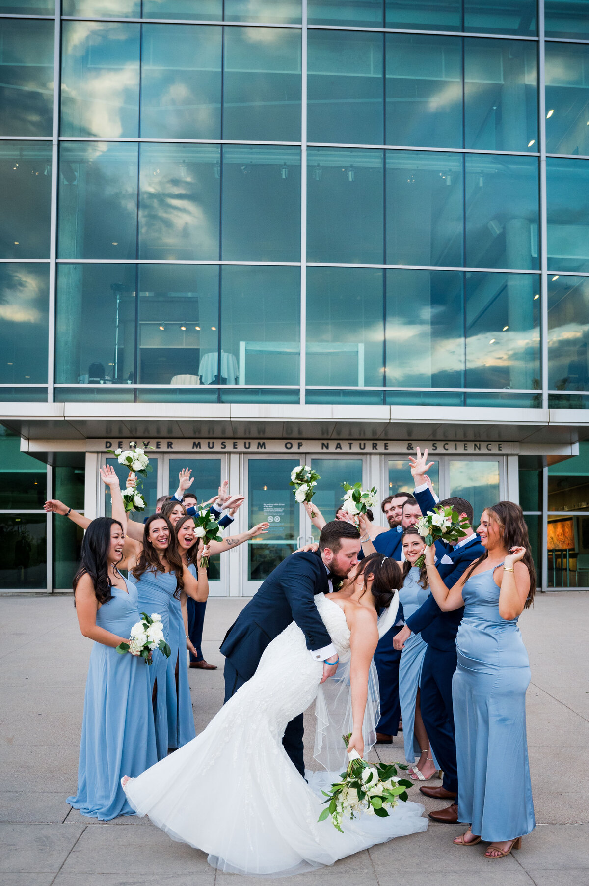A groom dips his bride for a kiss with half of the wedding party on one side and half on the other in front of The Denver Museum of Nature and Science.