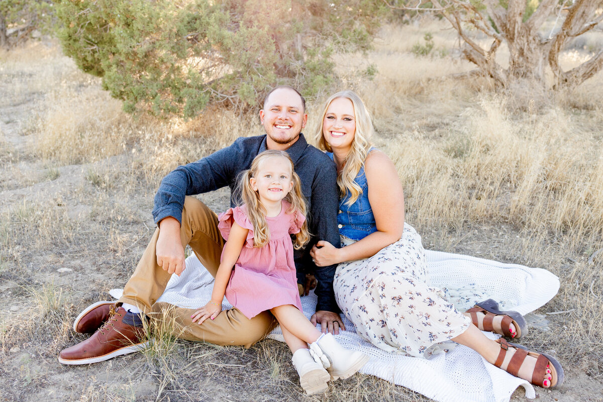 A smiling family sits together on a blanket in the dry, grassy fields of Eagle Mountain, Utah. The mother and father embrace their daughter, who beams at the camera.