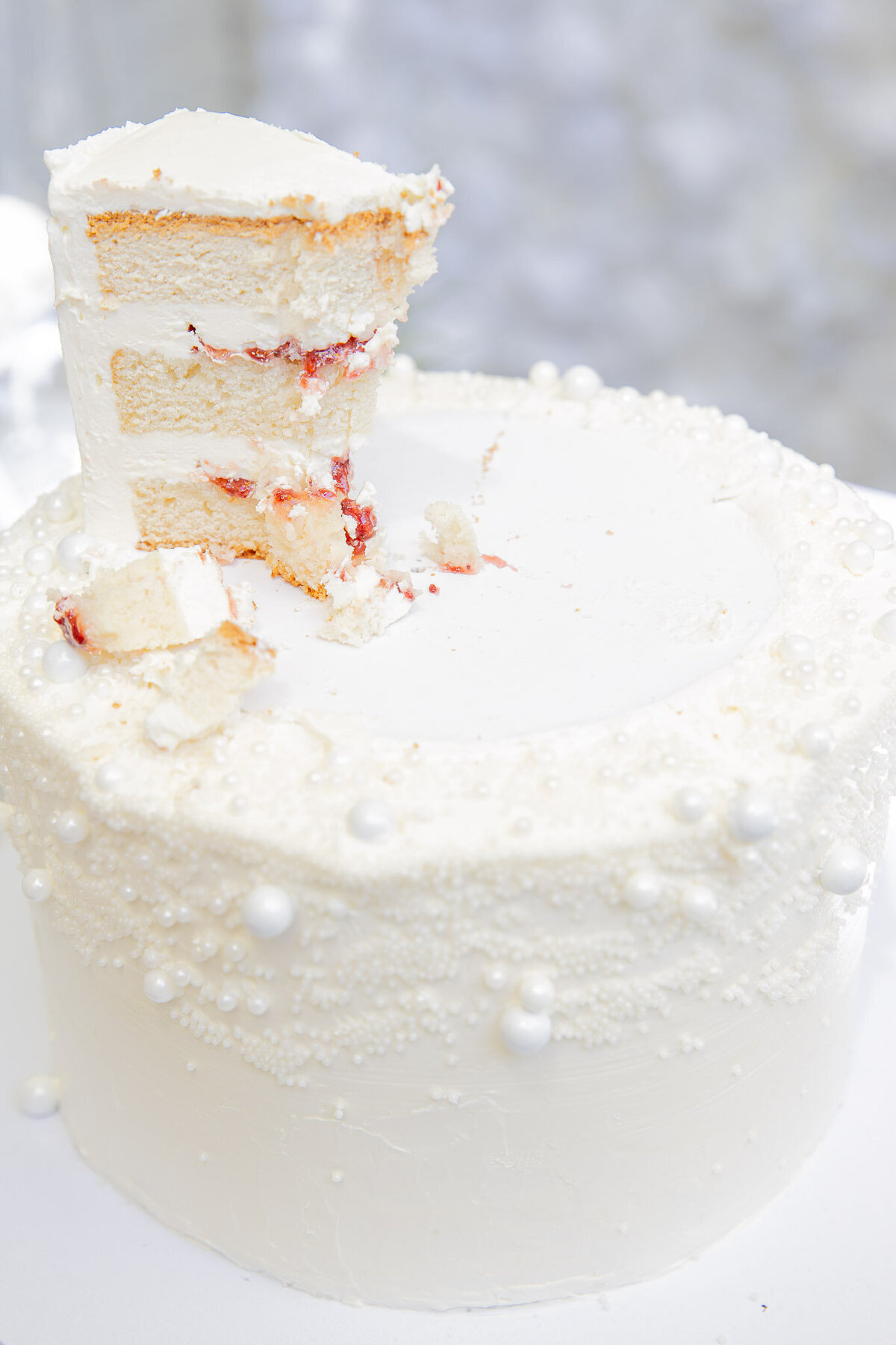 Close up of a wedding cake. The top layer is cut into with just one piece left. The piece of cake is a white cake with layers of white buttercream and berries.