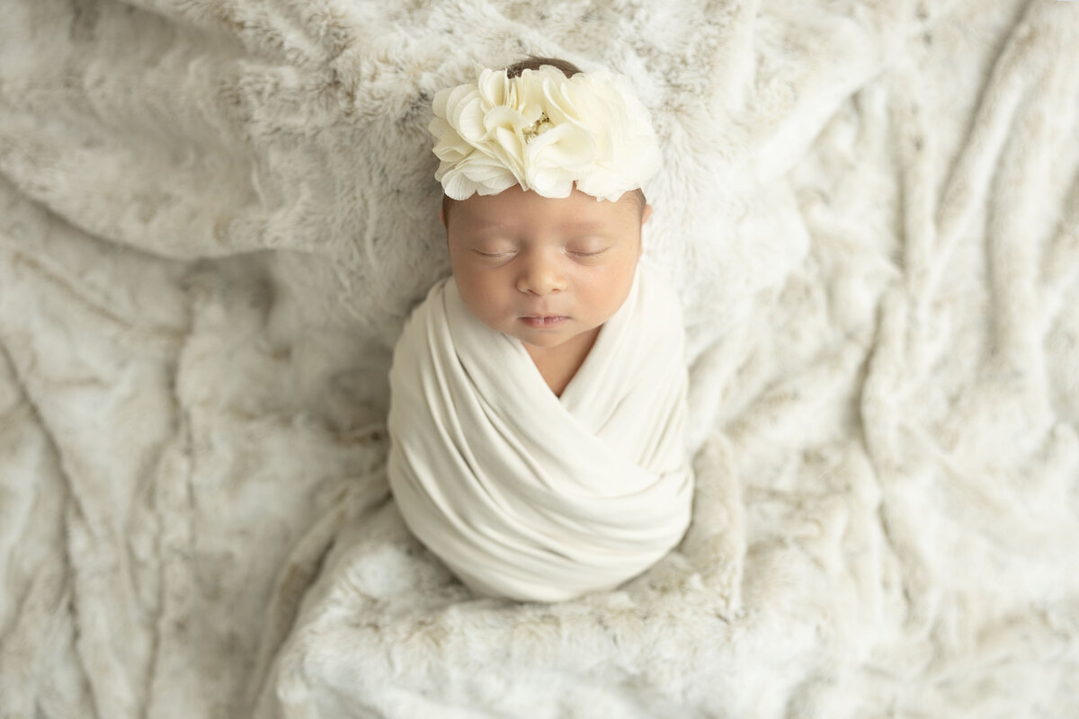 Baby lays back swaddled in a cream blanket with a flower headband.