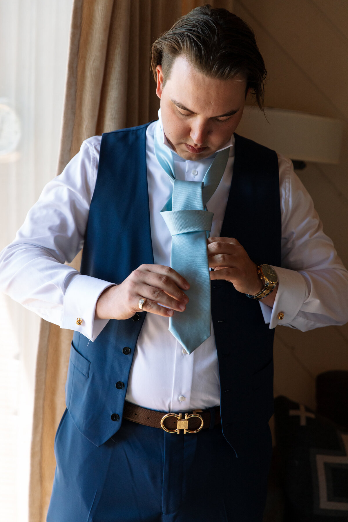 Groom in blue suit ties his tie near a window before his wedding.