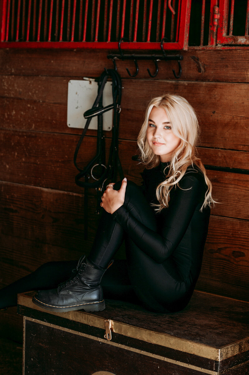 Senior girl in equestrian gear, seated in a rustic barn setting with tack and saddle.