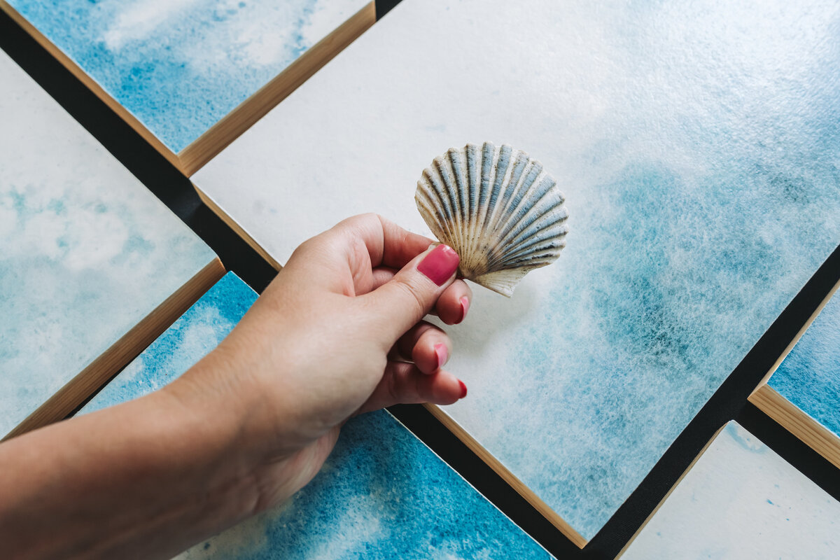 flat lay of watercolor paintings with woman's hand in front holding sea shell inspiration