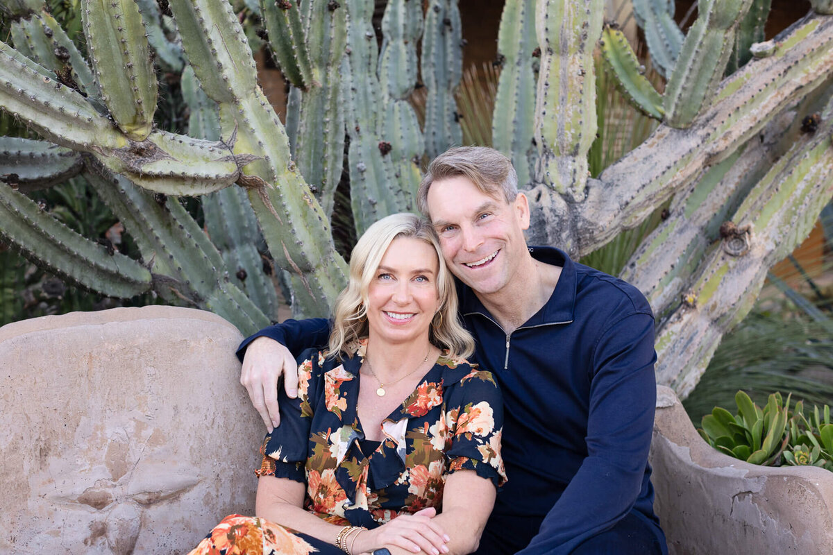 A husband wearing a navy, long sleeve shirt puts an arm around his blonde wife wearing a blue and orange dress as they both smile happily at the camera with a large cactus plant behind them.