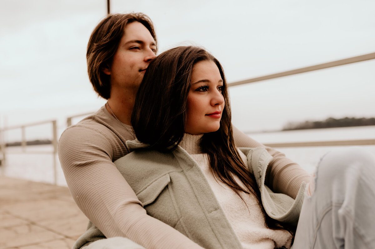 lake engagement photos with man and woman sitting on a dock together looking off to the distance while wearing neutral colors for their engagements
