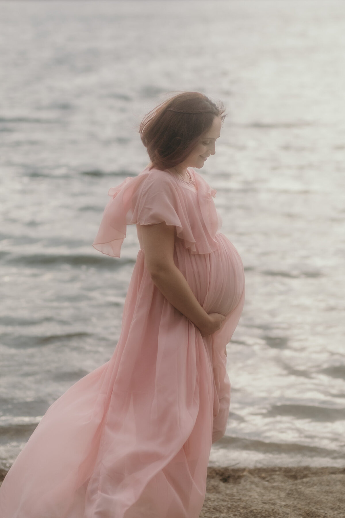 Pregnant woman in a flowing dress walks over sand at a sea in the sunlight.